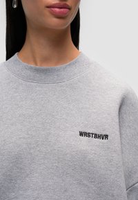 Felpa grigia con colletto tondo, caratterizzata da un piccolo logo nero "WRSTBHV" sul lato sinistro. Il tessuto sembra morbido e con una texture liscia.