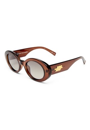 Lunettes de soleil ovales marron avec des verres dégradés et un logo géométrique doré sur la branche, présentées sur fond blanc.