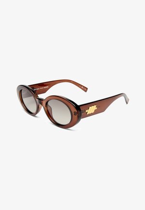 Lunettes de soleil ovales marron avec des verres dégradés et un logo géométrique doré sur la branche, présentées sur fond blanc.