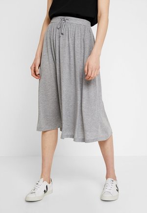 Femme portant une jupe grise ample longueur genou avec de fines rayures, un haut noir et des baskets blanches, debout devant un fond uni.