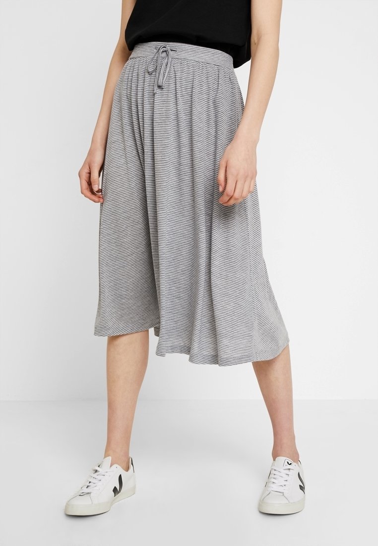 Femme portant une jupe grise ample longueur genou avec de fines rayures, un haut noir et des baskets blanches, debout devant un fond uni.