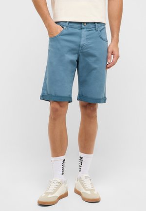 Mann trägt blaue Shorts mit aufgekrempeltem Saum, weiße Socken mit dem Schriftzug "Mustang" und beige Sneakers, steht vor einem einfarbigen hellen Hintergrund.