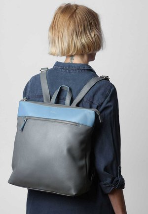 Zaino in pelle grigio con un accento azzurro chiaro, forma rettangolare, maniglie superiori e spallacci regolabili. Comparto con zip visibile.