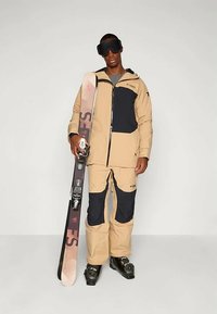 Skidress i beige og marineblå, laget av vanntett materiale. Har en glidelåsløs jakke, bukser, og svarte skistøvler. Holder ski.