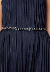 Ceinture chaîne métallique arborant un logo "MK", posée sur une robe en tissu plissé bleu marine. Le design de la chaîne présente une finition brillante.