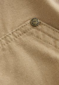 Tissu beige à surface texturée, avec un bouton en métal argenté embossé "Authentic 1862" et des coutures renforcées.