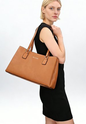 Blond vrouw in een mouwloze zwarte jurk met een grote beige leren tas over haar schouder tegen een witte achtergrond.