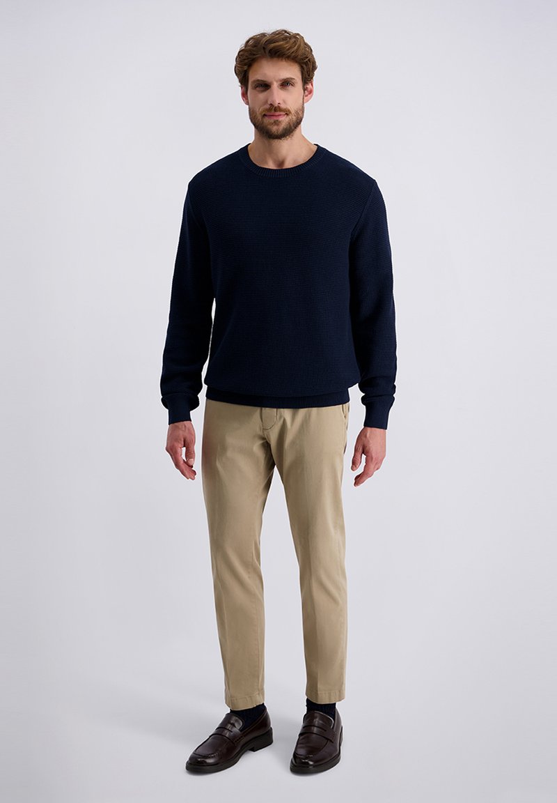 Donkerblauwe textuurtrui gecombineerd met beige broek en donkerbruine leren schoenen. De trui heeft een ronde halslijn en ribbels aan de mouwen.