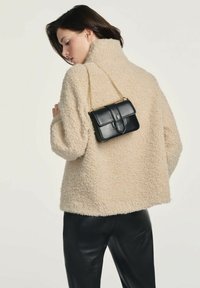 Beżowa, puszysta kurtka z polaru o wysokim kołnierzu, noszona na czarnych skórzanych spodniach. Mała czarna skórzana torba wisi na złotym łańcuszku.