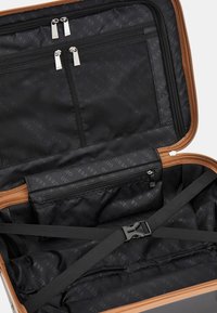 Valigia rigida con accenti marroni; l'interno presenta una fodera nera con il logo "GUESS" ripetuto, tasche con zip e cinghie regolabili.