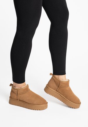 Person iført sorte leggings og lysebrune ankelstøvler i ruskind med foret inderside og tykke gummisåler på hvid baggrund.