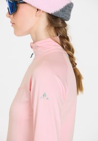Helles rosa Langarmoberteil mit halbem Reißverschluss am Kragen und reflektierendem Logo am Ärmel. Glatte Textur, taillierte Passform und gefaltete graue Strickmütze.