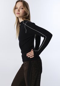 Jeune femme aux longs cheveux blonds posant dans un pull en rib de couleur noire avec des bordures blanches et un pantalon brun foncé avec un panneau à motif léopard.