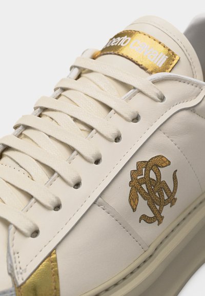 Κοντινή λήψη λευκού δερμάτινου αθλητικού παπουτσιού με μπεζ κορδόνια, χρυσό μεταλλικό λογότυπο στη γλώσσα και έμβλημα φιδιού στην πλευρική πάνελ.