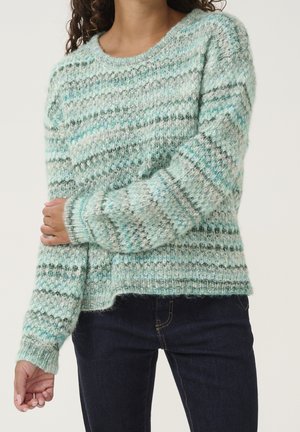 Femme portant un pull en maille duveteuse à rayures horizontales dans des tons de bleu, gris et blanc, associé à un jean foncé.
