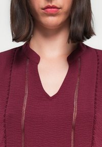 Blouse bordeaux en tissu texturé, décolleté en V et accents en dentelle sur les côtés, dotée d'une coupe décontractée et de détails subtils.