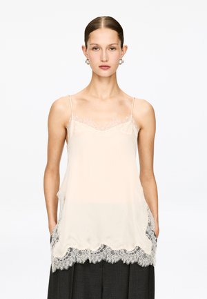 Cremefarbenes Camisole mit Spitzenbesatz und dünnen Trägern, ausgestattet mit einem gezackten Saum und glatter Satinstruktur. Über einem dunklen Kleidungsstück getragen.