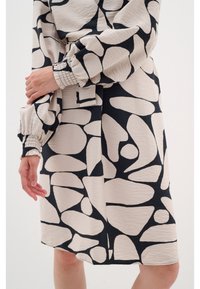 Robe abstraite noire et beige à motifs, longueur genou, avec de longues manches bouffantes et des poignets élastiques, portée par une personne sur un fond uni.