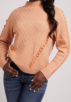 Brzoskwiniowy sweter w splot kablowy z skórzanymi wstążkami wzdłuż rękawów i przodu, noszony z ciemnoniebieskimi dżinsami, dłonie spoczywają na biodrach.