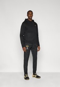 Μαύρο φούτερ με κουκούλα και τσέπη καγκουρό, μαύρο παντελόνι στενής γραμμής με γεωμετρικό μοτίβο και μαύρα παπούτσια με μπεζ λεπτομέρειες. Χρυσές λεπτομέρειες λογότυπου.