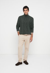 Homem com suéter de gola alta em malha grossa verde escuro, calças bege, mocassins castanhos, de pé com as mãos nos bolsos contra um fundo simples.