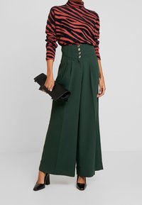 Pantaloni larghi verdi con vita alta e pieghe, dotati di bottoni dorati. Abbinati a un maglione a righe nero e rosa e una pochette nera.