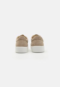 Beige semskinn sneakers med rund hæl og hvit gummisåle. Har polstret krage og bakre trekkstropp. Minimal merkevare på hælen.