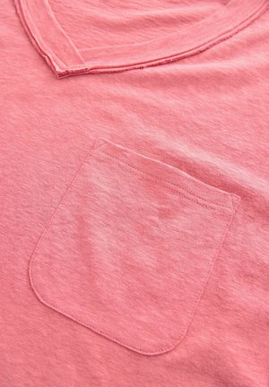 Ljusrosa T-shirt i mjukt tyg, med rundad halsringning och en bröstficka med en kurvad övre kant. Subtil struktur synlig.
