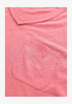 T-shirt rose pâle en tissu doux, dotée d'un col rond et d'une poche poitrine avec un bord supérieur arrondi. Texture subtile visible.