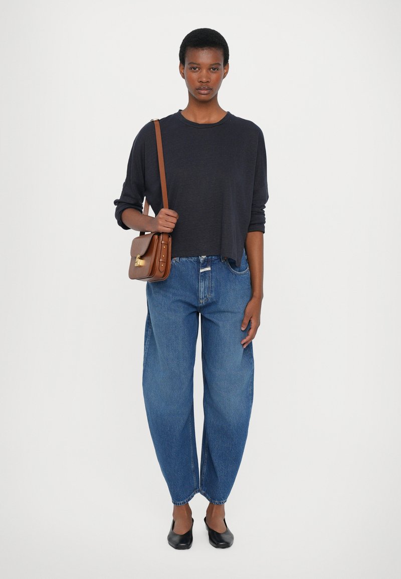 Navyblaues Langarmoberteil, lockere Passform, mit hochgeschnittenen hellblauen Jeans. Braune Umhängetasche mit goldenen Details und schwarze flache Schuhe.