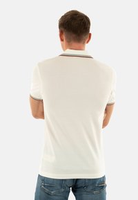 Hvid polo-skjorte med korte ærmer, der har en ribbet krave med kontraststriber. Modellen bærer blå jeans, hvor skjortens tekstur er synlig.