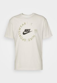 T-shirt en coton crème avec un grand logo Nike noir et un texte circulaire vert indiquant "Sportswear" et "Depuis 1972". Encolure classique.