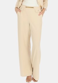 Beżowe spodnie o szerokich nogawkach z gładką teksturą, z mosiężnym łańcuchem na pasku i bocznymi kieszeniami.