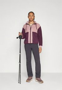 Bordeauxrode en roze fleecejas met een ritssluiting aan de voorkant en capuchon, gecombineerd met een donkere broek en wandelstokken. De jas heeft geometrische kleurvlakken.