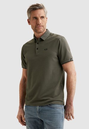 Mann mit grauen Haaren und Bart, trägt ein olivgrünes Poloshirt und hellblaue Jeans, schaut zur Seite vor einem einfarbigen Hintergrund.