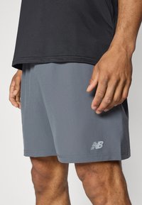 Man som bär grå träningsshorts med New Balance-logga nära fållen och svart tröja, stående med händerna avslappnat vid sidorna.