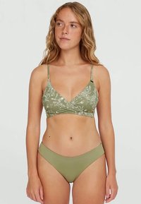 Νεαρή γυναίκα με κυματιστά ξανθά μαλλιά, φορώντας ελαιόχρωμο μπικίνι με floral εκτύπωση στην κορυφή και απλή κάτω μεριά, στραμμένη μπροστά σε απλό φόντο.