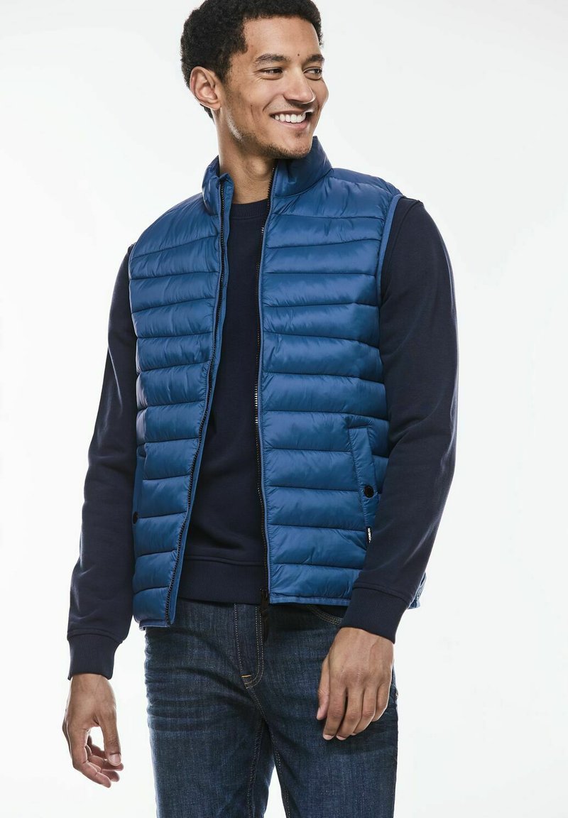 Modrá vycpaná vesta s vysokým límcem, zapínáním na zip a bočními kapsami, nošená přes námořnický svetr. Neformální design, hladká textura, lehký vzhled.