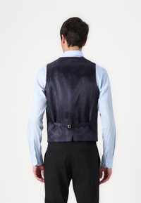 Colete azul marinho com textura padrão, design slim fit, alça traseira ajustável única, combinado com uma camisa dress azul clara e calças pretas.