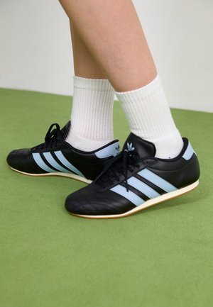 Nogi w białych prążkowanych skarpetach za kostkę i czarnych butach sportowych Adidas z jasnoniebieskimi paskami stojące na zielonej podłodze.