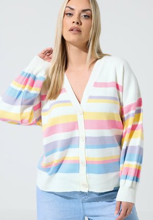 Lyshåret kvinde iført en hvid cardigan med pink, gul, blå og lilla vandrette striber, kombineret med lyseblå jeans, mod hvid baggrund.
