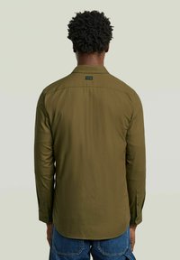 Homme avec des cheveux courts et bouclés portant une chemise à manches longues vert olive et un jean en denim bleu, se tenant de dos contre un fond uni.