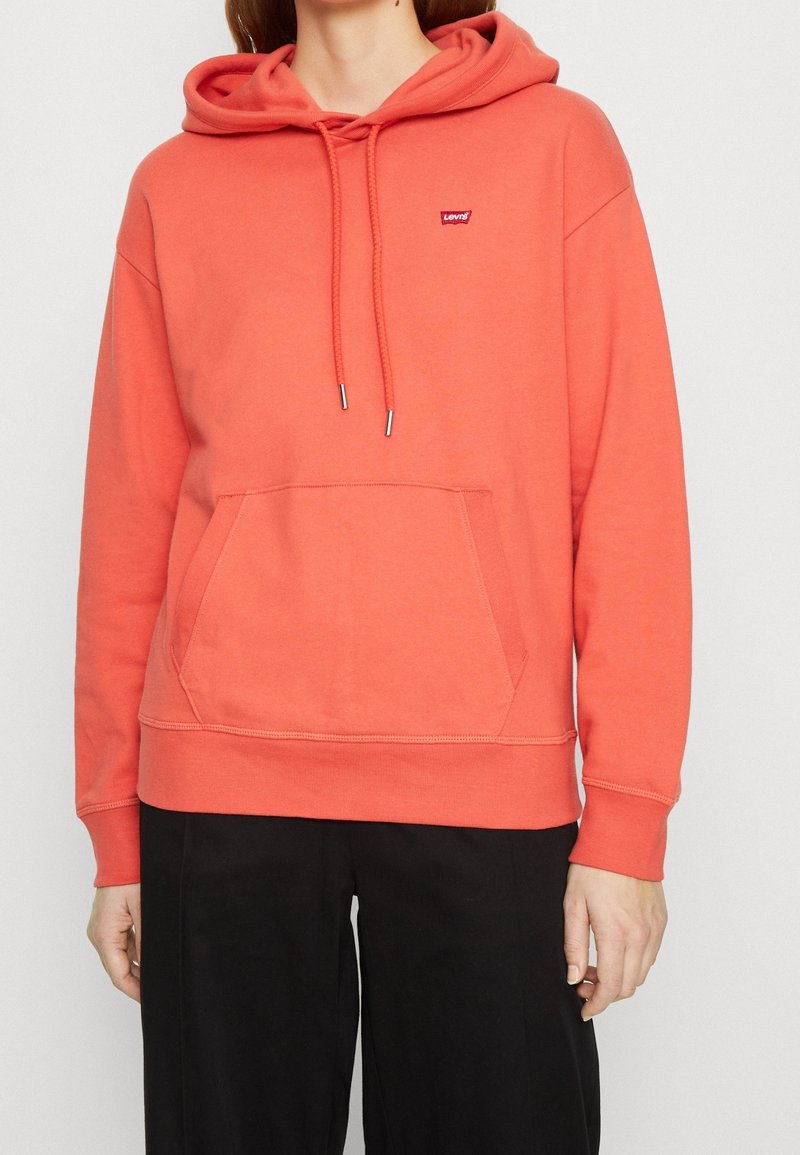 Coral hoodie made of soft fabric featuring a front pocket, drawstring hood, and a small logo patch on the chest. Classic pullover design.