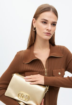 Un mannequin portant un pull côtelé marron tient un sac à main doré brillant, arborant un logo, sur un fond blanc uni.