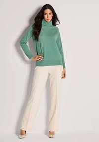 Hellgrüner Rollkragenpullover mit langen Ärmeln, kombiniert mit cremefarbenen Weitbein-Hosen und gemusterten Absätzen, vor einem neutralen Hintergrund.