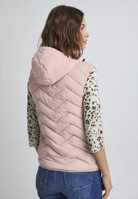 Gilet en duvet rose clair avec une capuche, présentant un motif en chevrons sur le dos, associé à une chemise à manches longues crème avec un imprimé floral.