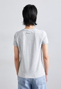 Tricou scurt gri cu mânecă scurtă și guler rotund, având o etichetă mică la spate și o textură netedă și moale. Purtați-l cu blugi de culoare albastru deschis.