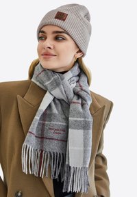 Grå och vit rutad halsduk med fransar, med röd text "Victoria Hyde London," bärs tillsammans med en beige ribbad stickad mössa.