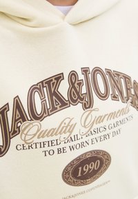 Hoodie crème avec un grand texte marron qui dit "JACK & JONES" et un texte plus petit en dessous. Comprend un graphique circulaire avec "1990". Texture de tissu douce.
