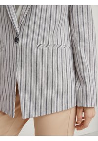 Blazer a rayas de tela ligera con rayas verticales en azul y blanco. Cuenta con un solo botón, solapas de muesca y dos bolsillos frontales.
